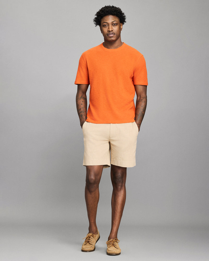 Man wearing an orange t-shirt and beige shorts against a gray background Alternate