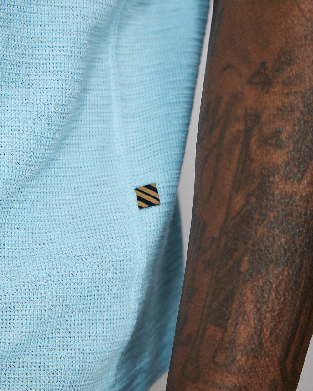 Light blue textured fabric with a small black and yellow striped logo, next to a tattooed arm.