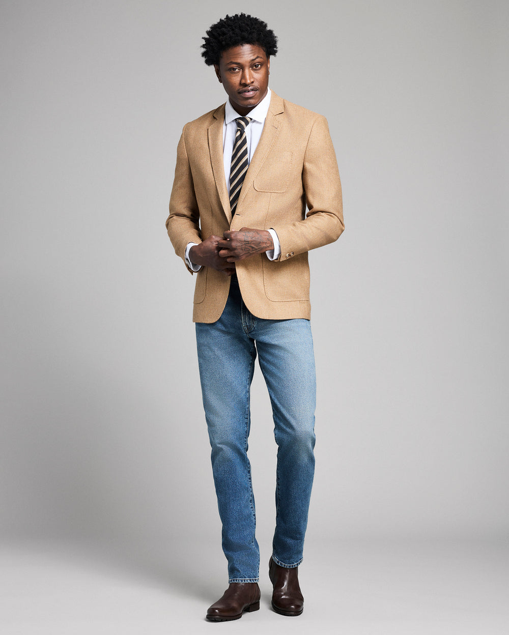 Man wearing a beige blazer, blue jeans, and a striped tie on a gray background