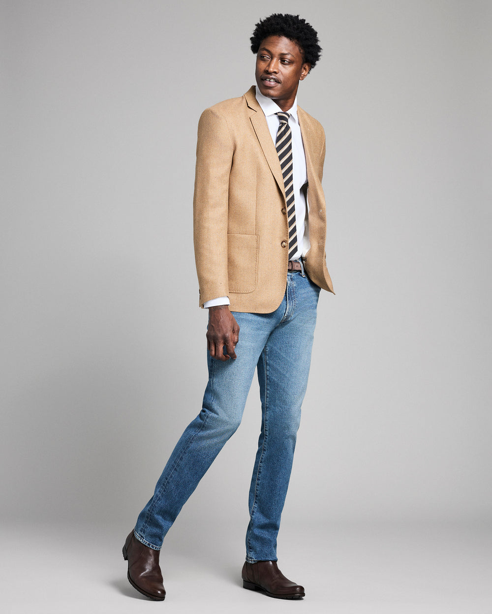 Man wearing a beige blazer, striped tie, and blue jeans on a gray background