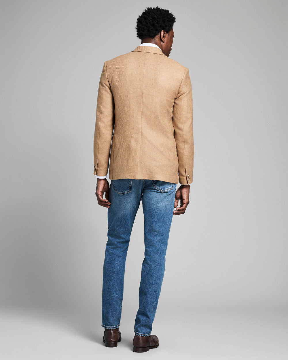 Man wearing a beige blazer and blue jeans on a gray background