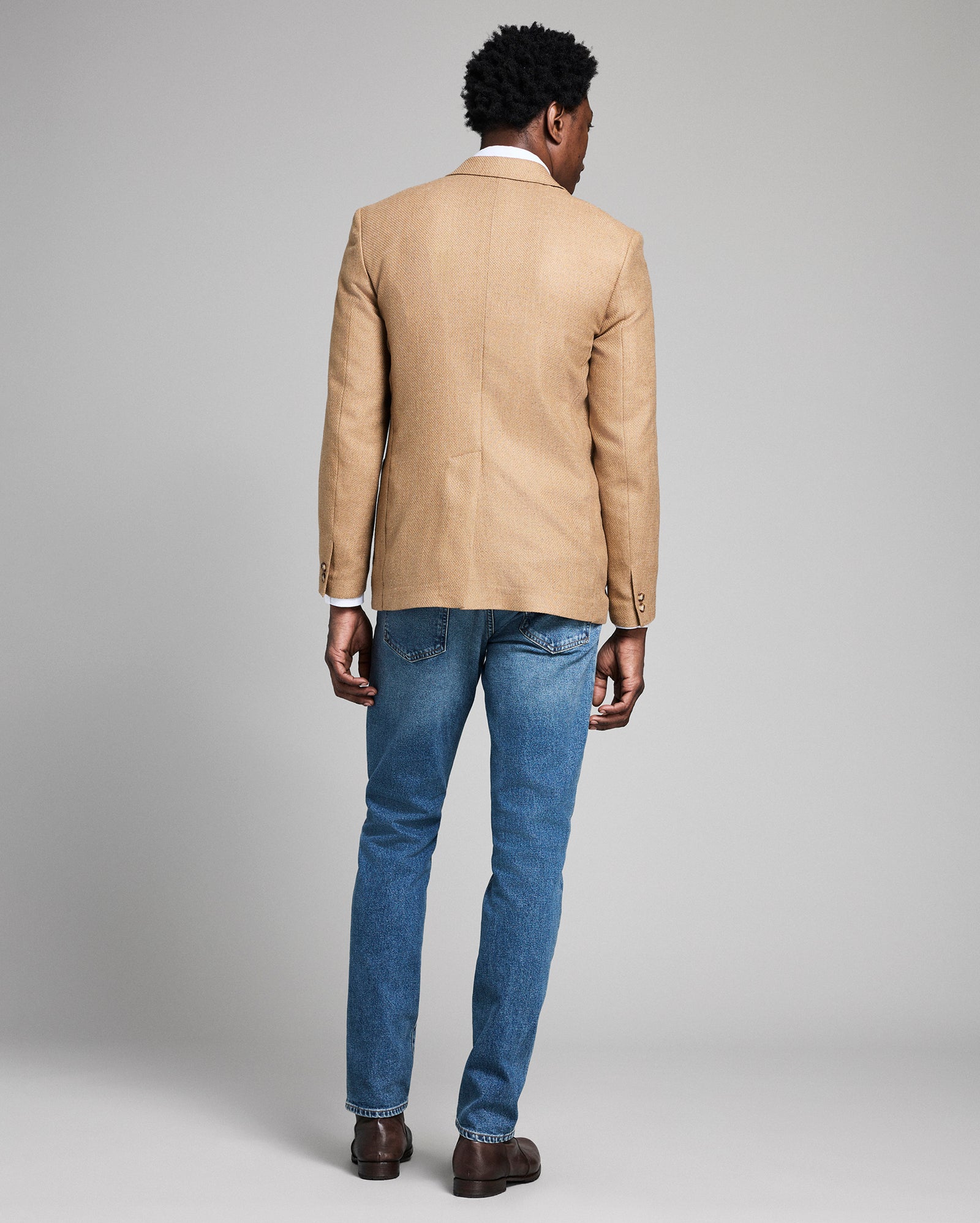 Man wearing a beige blazer and blue jeans on a gray background