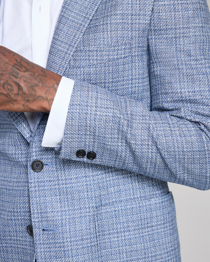 Close-up of a blue textured blazer with a white shirt and tattooed arm.