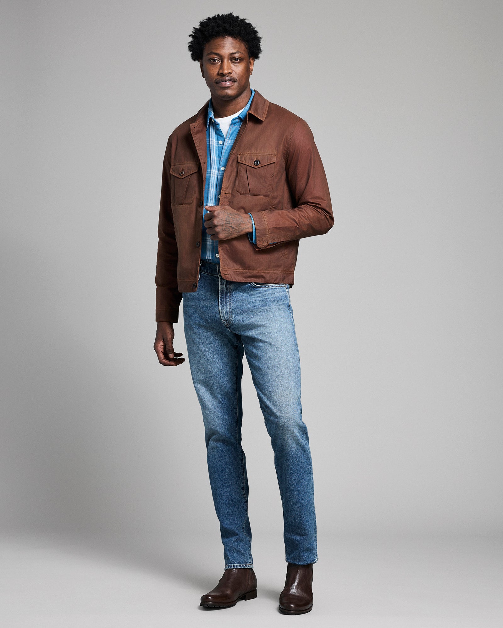 Man wearing a brown jacket, blue shirt, and jeans on a gray background