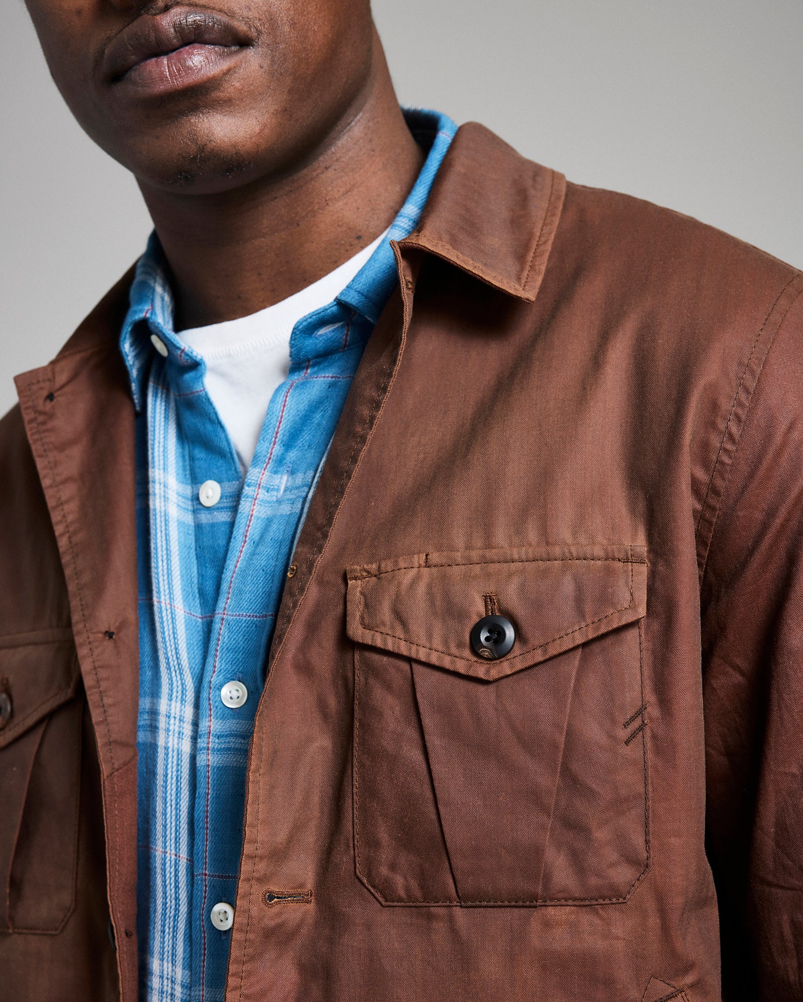 Man wearing a brown jacket over a blue plaid shirt with a neutral background