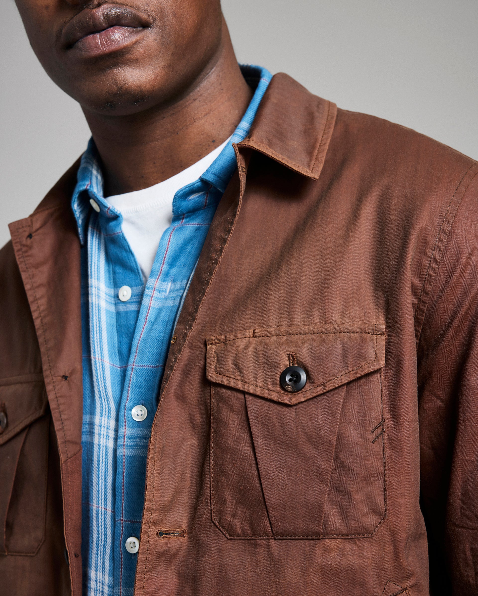 Man wearing a brown jacket over a blue plaid shirt with a neutral background