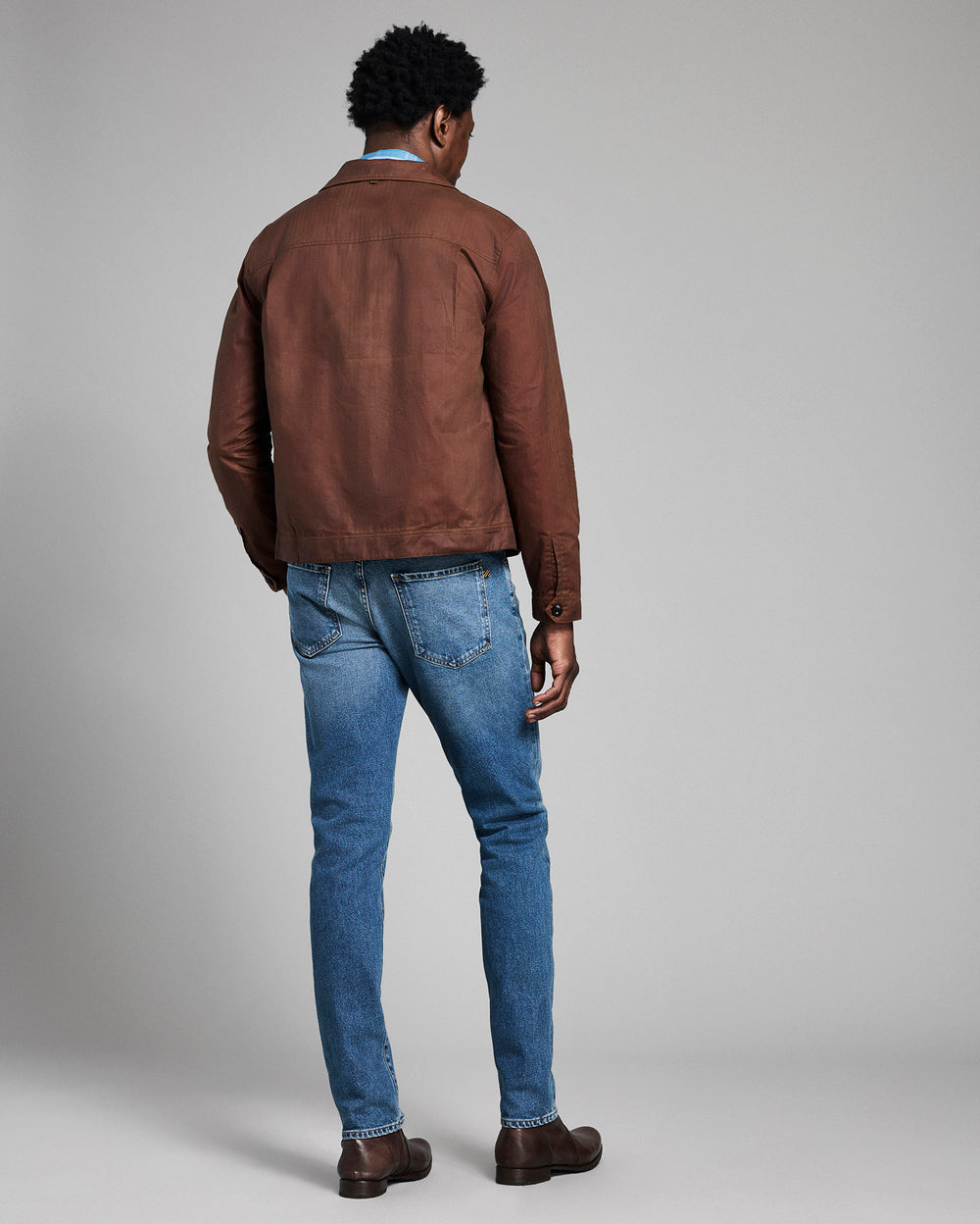 Man wearing a brown leather jacket and blue jeans against a gray background