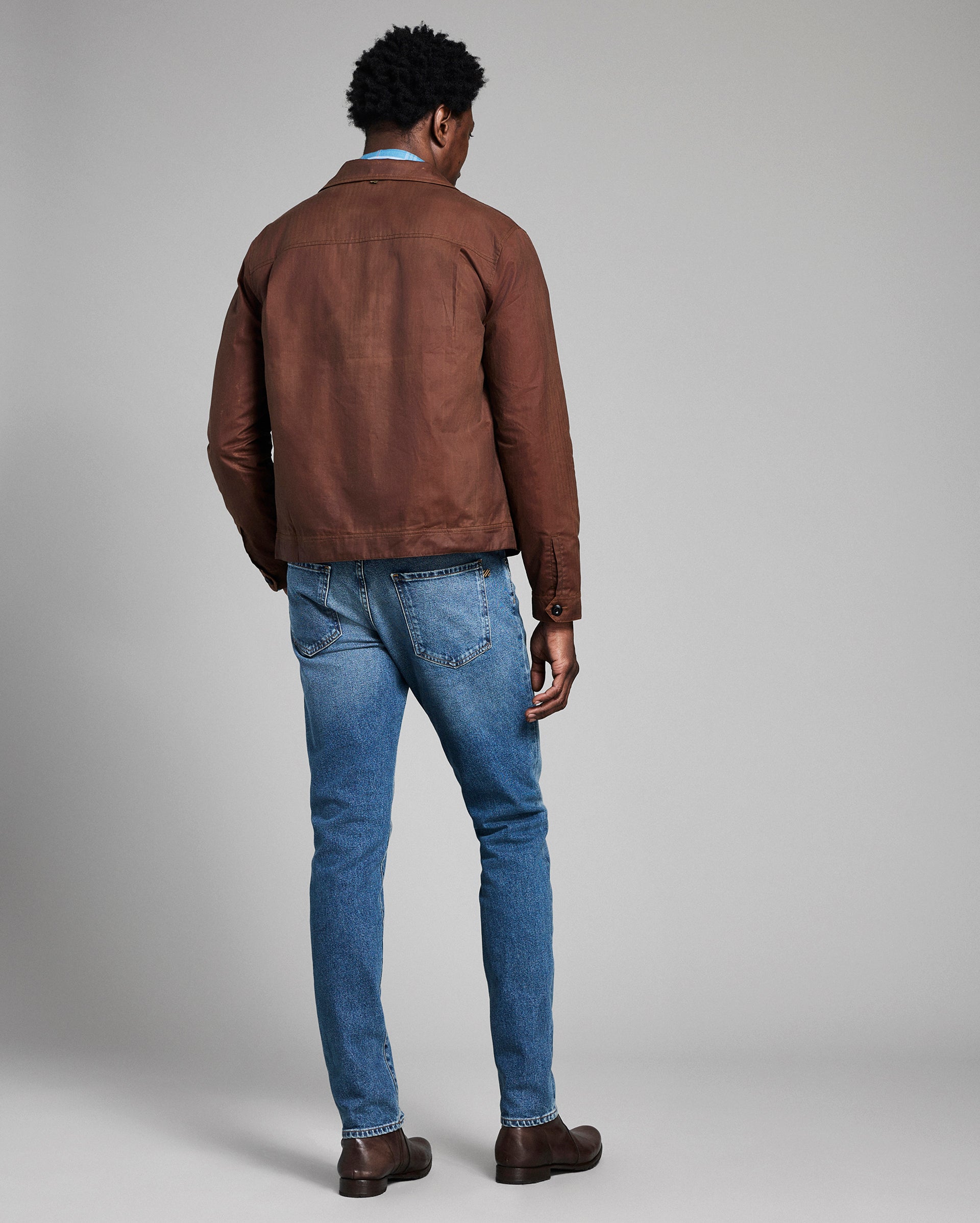 Man wearing a brown leather jacket and blue jeans against a gray background