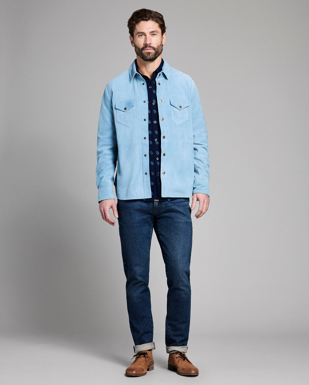 Man wearing a light blue denim jacket over a dark shirt, paired with dark jeans and brown shoes on a gray background.