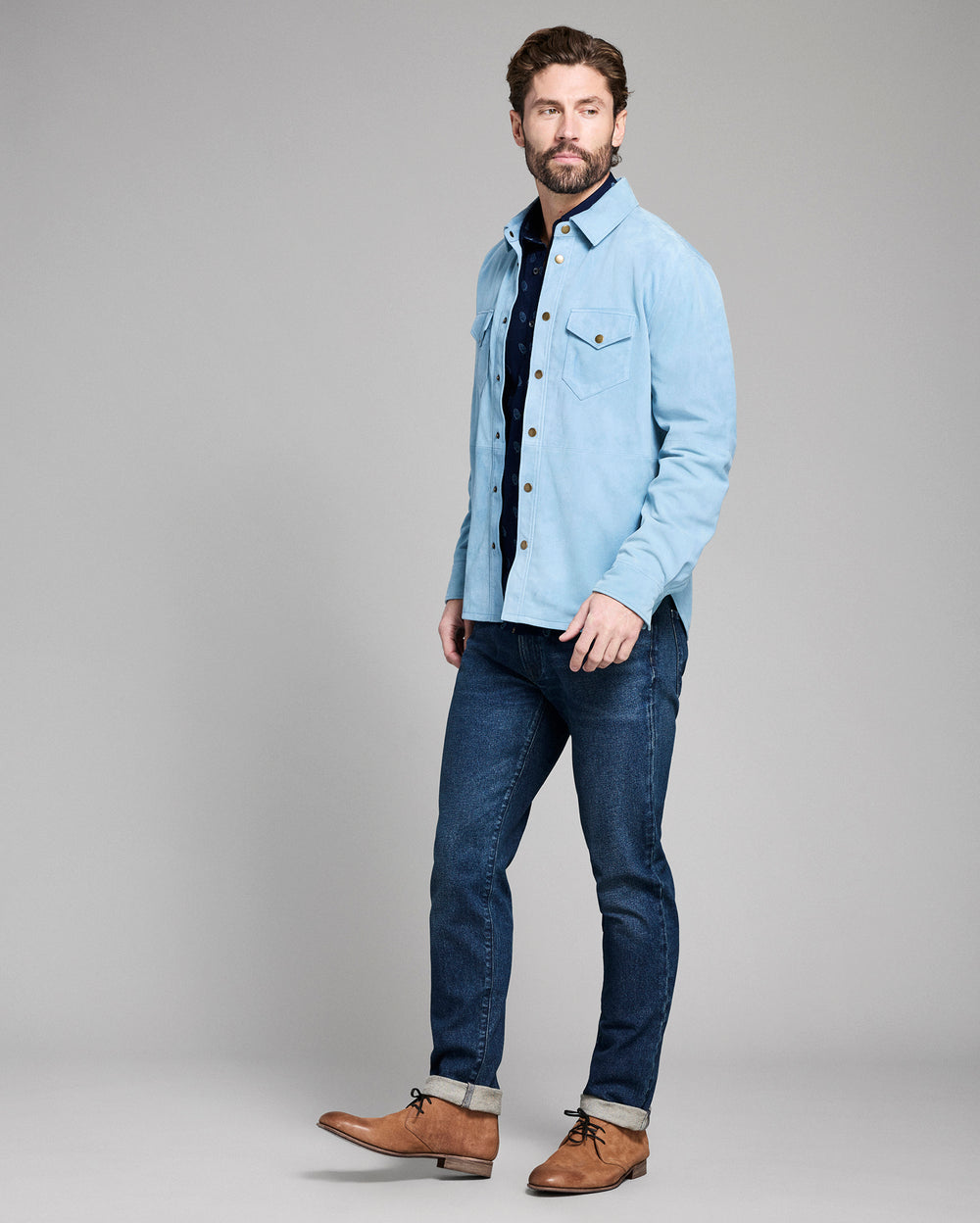 Man wearing a light blue denim shirt, dark jeans, and brown shoes on a gray background