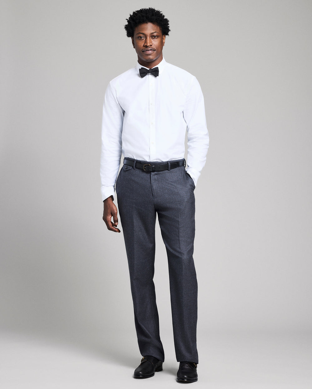 Man wearing a white dress shirt, black bow tie, dark gray pants, and black shoes on a plain background