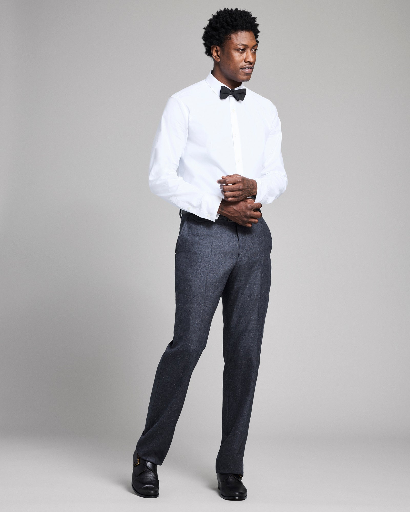 Man wearing a white dress shirt, black bow tie, gray pants, and black shoes on a plain background