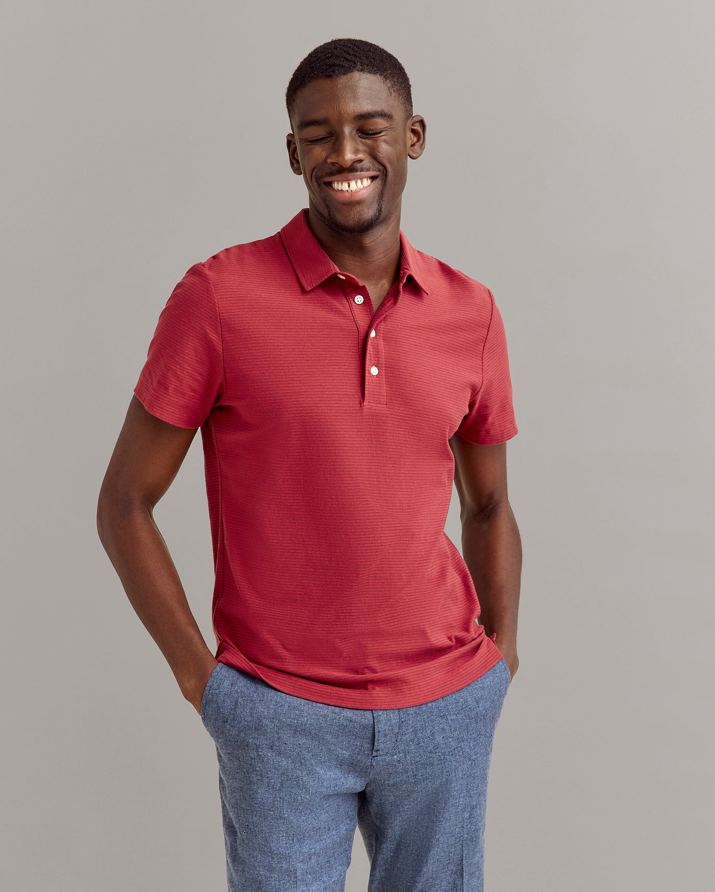 A man smiles with his eyes closed, wearing a short-sleeved red polo shirt and blue pants. His hands are in his pockets, and he stands against a plain light gray background.