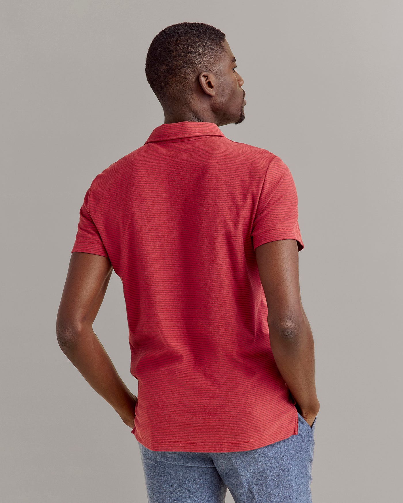 A man stands facing away, wearing a short-sleeve red polo shirt and blue pants, with his hands in his pockets against a plain, light-colored background.