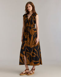Woman wearing a sleeveless, black and brown floral maxi dress with a v-neck and front slit, paired with tan wedge sandals, standing against a plain light background.