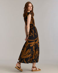 A woman with light brown hair wears a black and brown patterned maxi dress and tan sandals, standing sideways and looking over her shoulder against a plain light background.