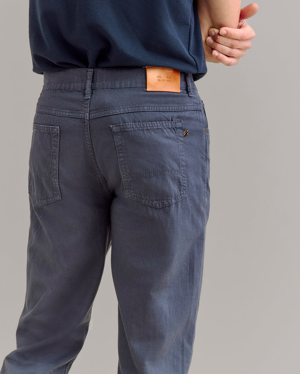 A person wearing blue pants and a dark blue shirt stands with one arm behind their back. The focus is on the back of the pants, which have patch pockets and a tan leather label on the waistband.