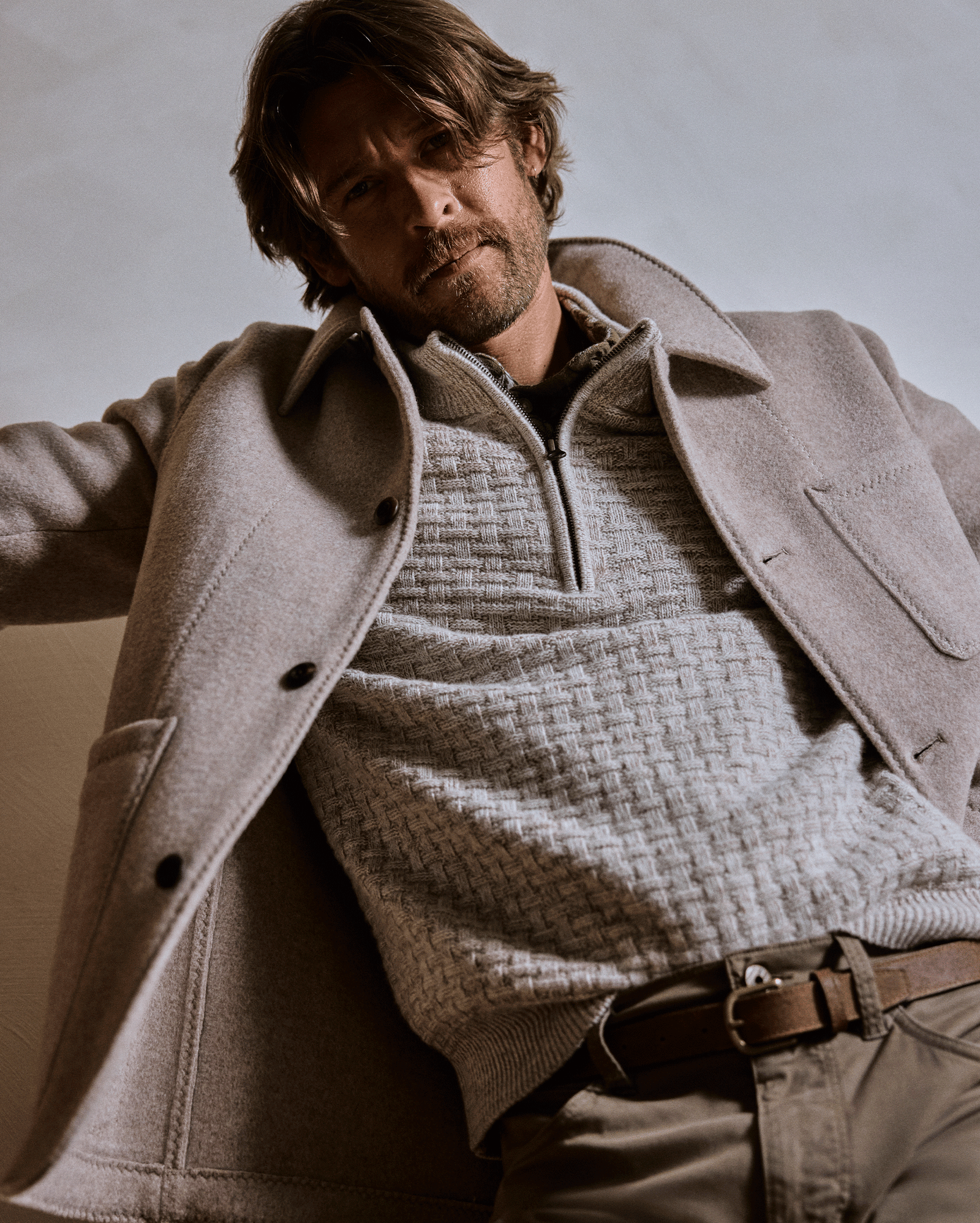 Man wearing a beige coat over a textured sweater against a neutral background