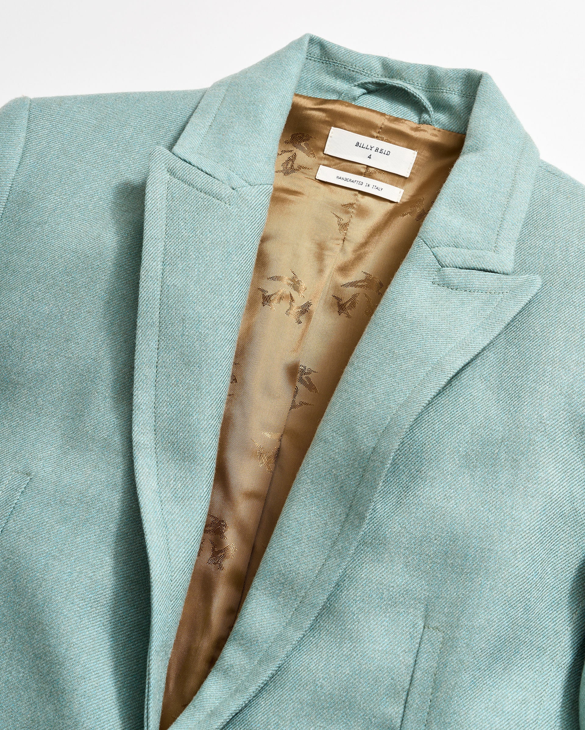 A close-up of a light teal blazer with a brown silky inner lining that has a subtle floral pattern. The inside label on the collar reads MADEWELL and MADE IN ITALY.
