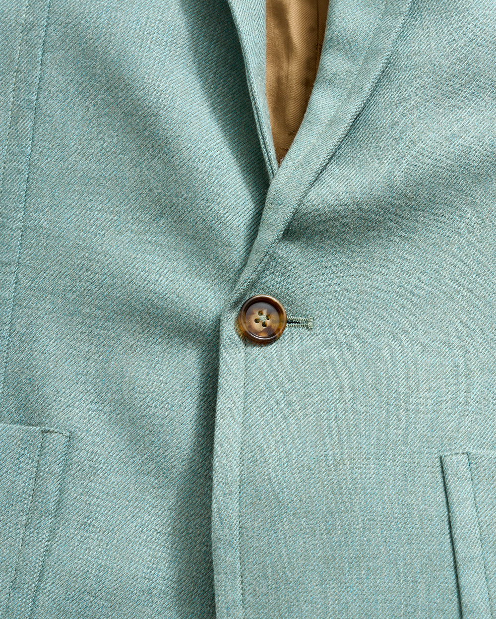 Close-up of a light teal blazer with a single brown button fastened. The blazer features a notched lapel and two front pockets, with a glimpse of the tan inner lining visible near the collar.