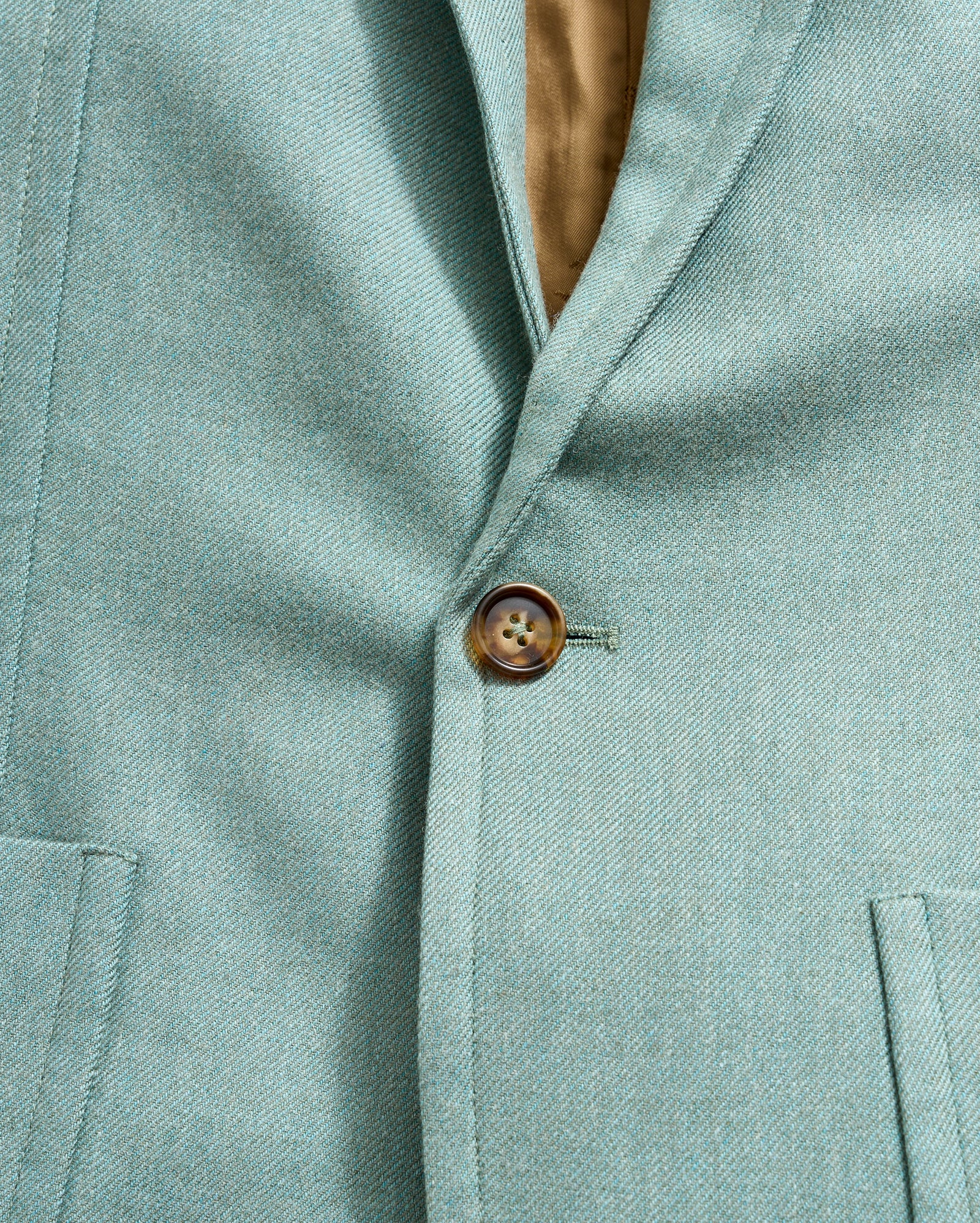 Close-up of a light teal blazer with a single brown button fastened. The blazer features a notched lapel and two front pockets, with a glimpse of the tan inner lining visible near the collar.
