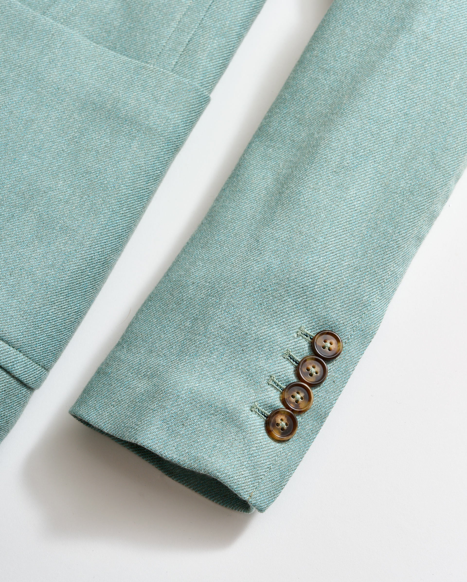 A close-up of a light teal blazer sleeve with four brown buttons along the cuff, lying flat on a white surface. The jacket pocket and sleeve seam are also visible.