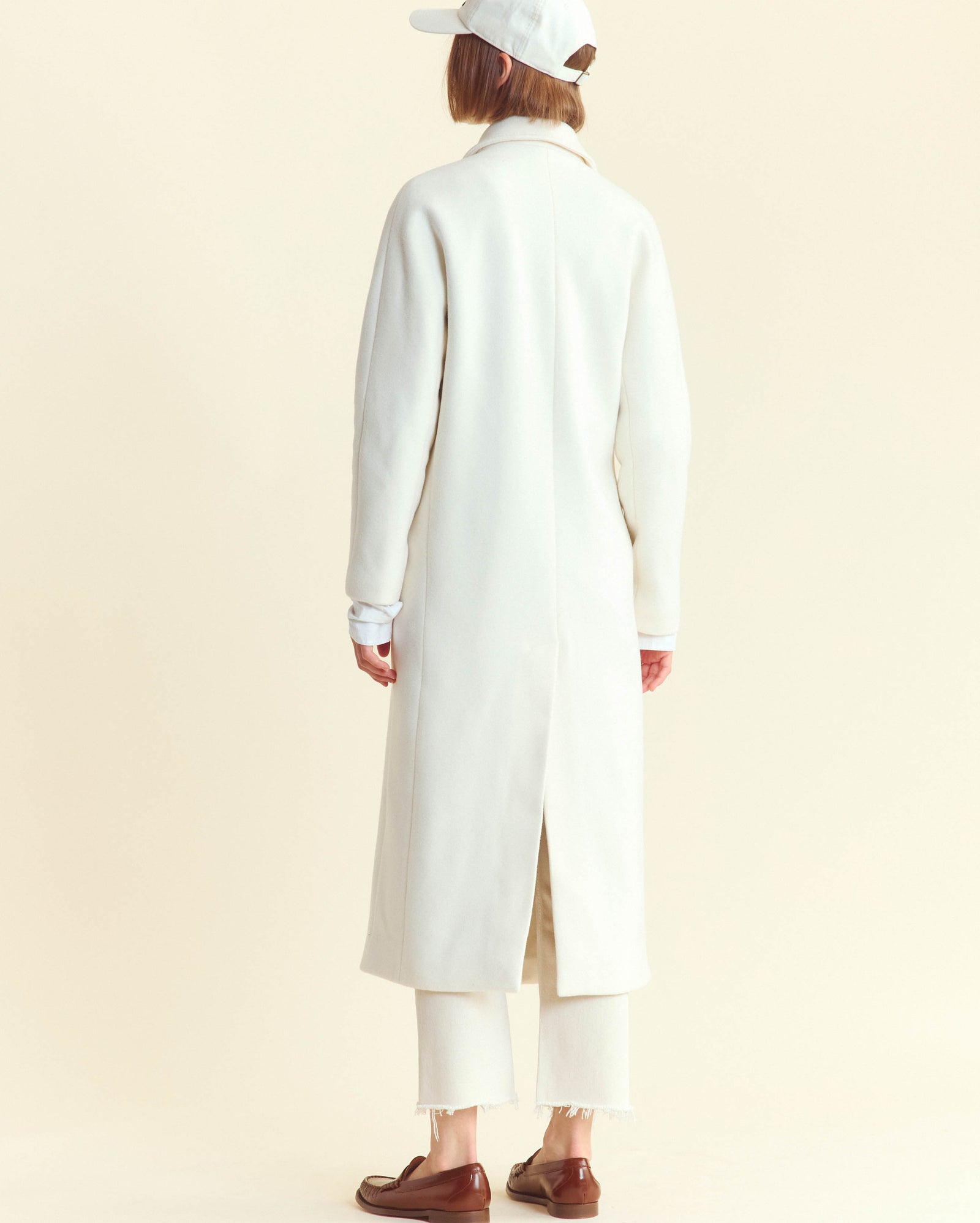 A person stands facing away, wearing a long white coat, white cropped pants, white cap, and brown loafers. The background is a plain light color.