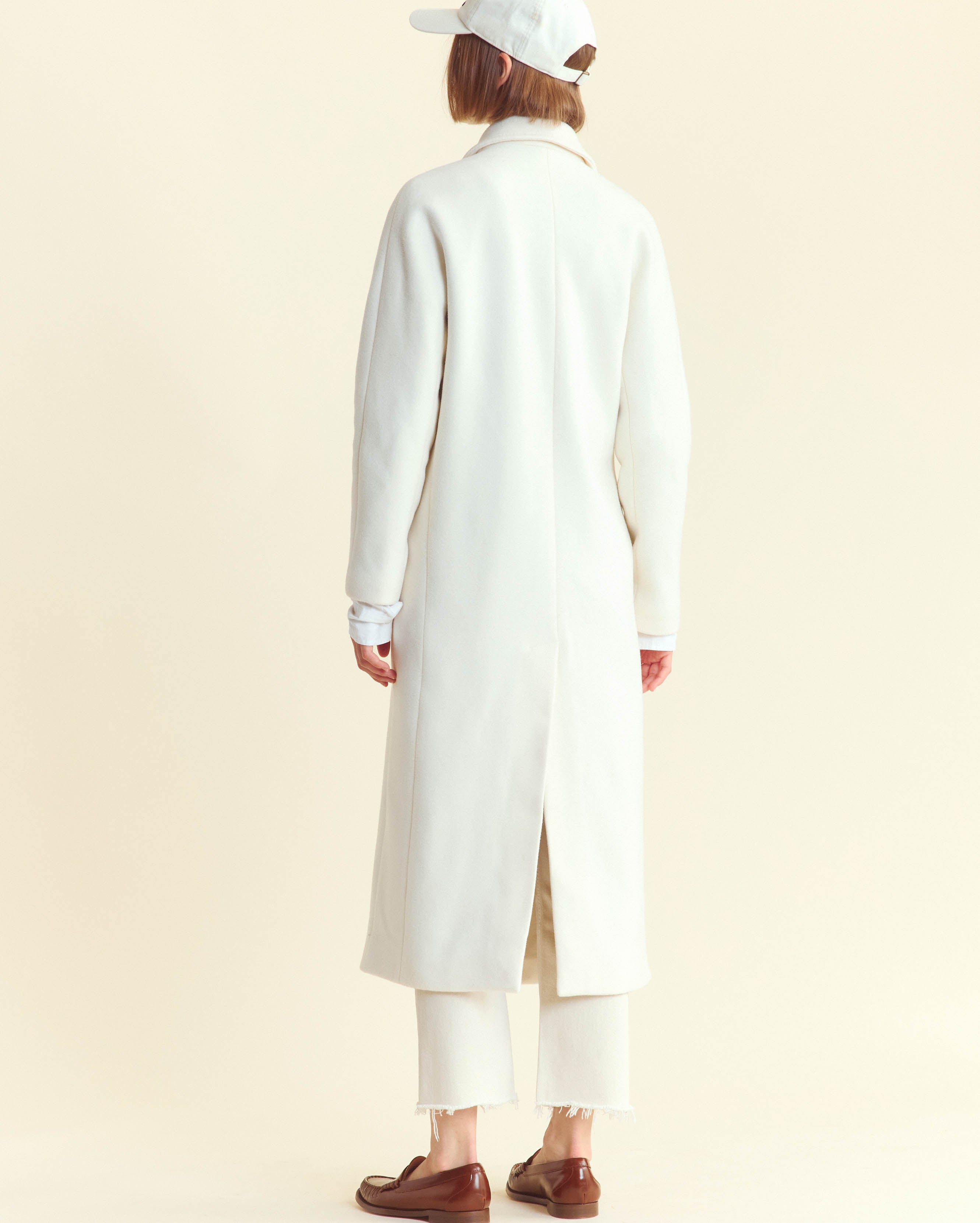 A person stands facing away, wearing a long white coat, white cropped pants, white cap, and brown loafers. The background is a plain light color.