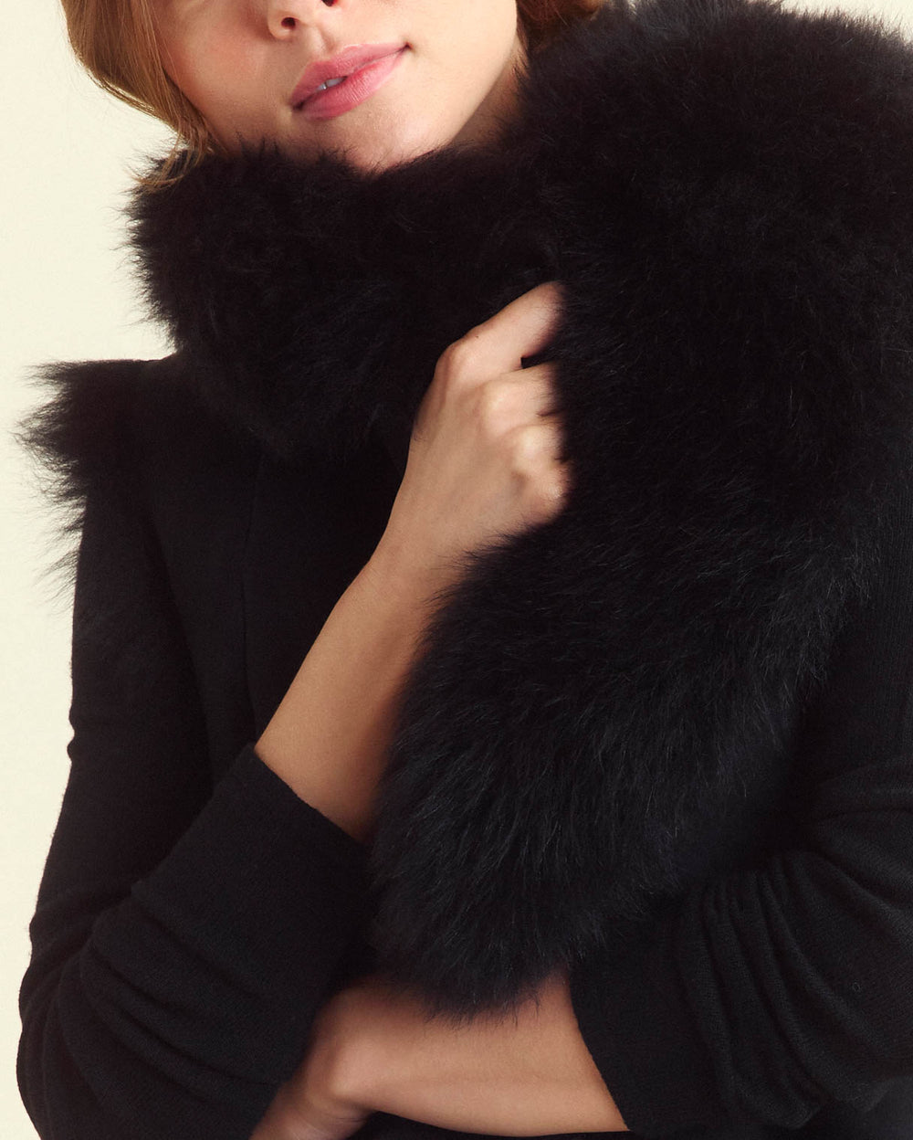 A person wearing black holds a fluffy black fur scarf close to their chest, partially covering their face and shoulders.