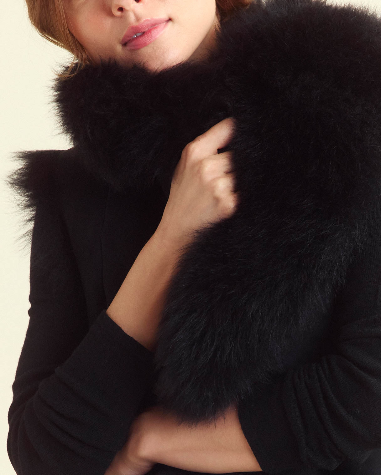 A person wearing black holds a fluffy black fur scarf close to their chest, partially covering their face and shoulders.