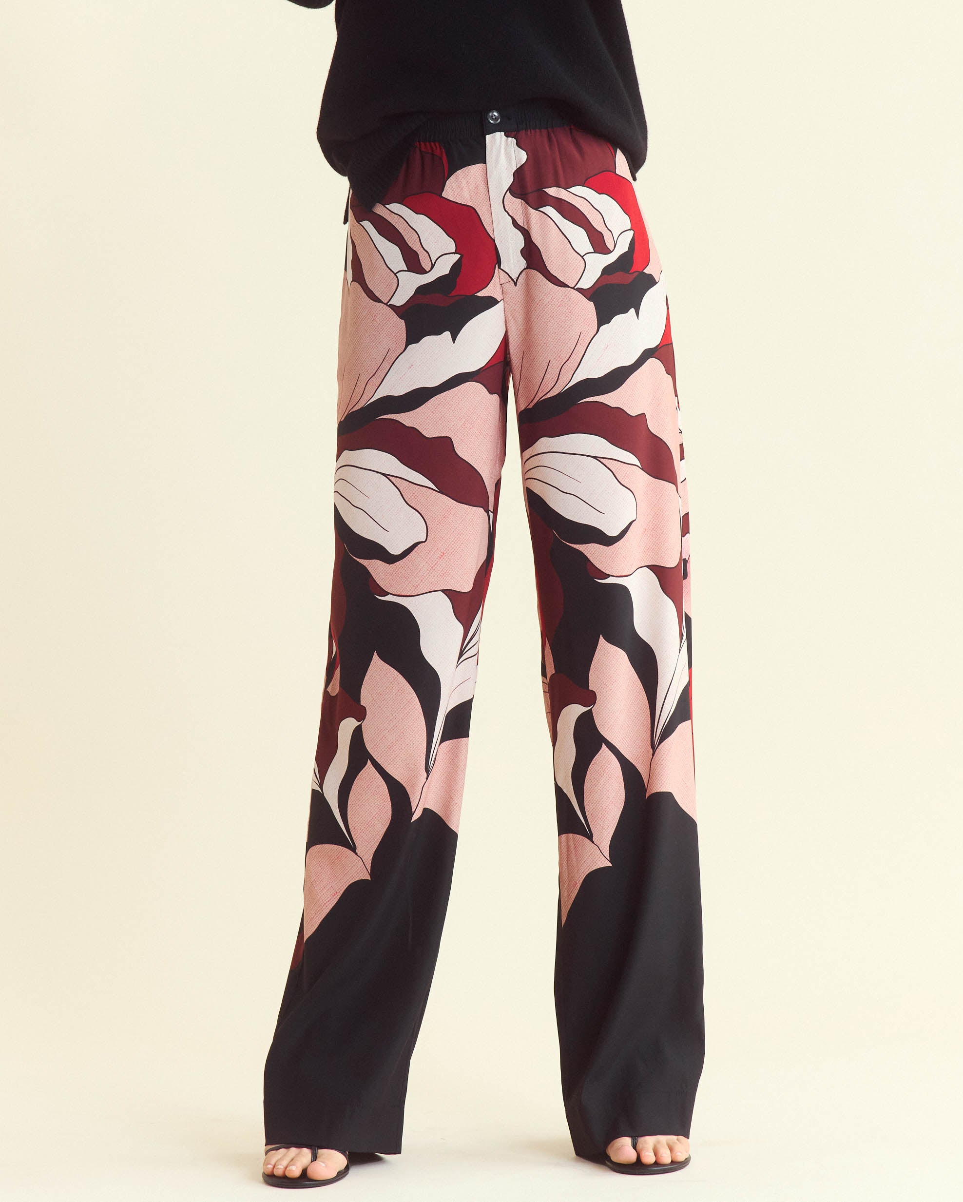 Person wearing wide-leg pants with a bold, abstract pink, red, black, and white floral pattern. Only the lower half of the body is visible; feet are in sandals and the background is light beige.