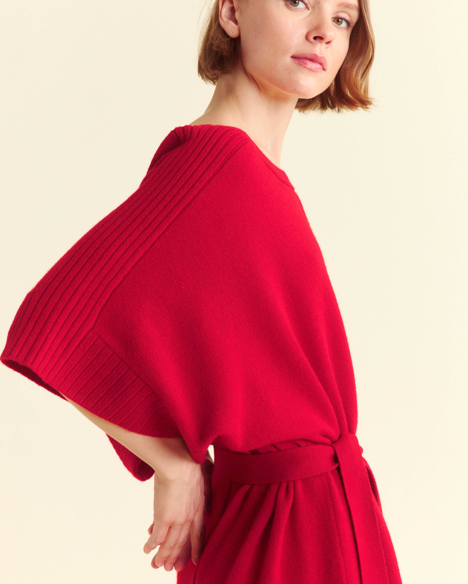 A woman with short light brown hair wears a loose-fitting, bright red dress with wide sleeves and a cinched waist, posing against a neutral background.