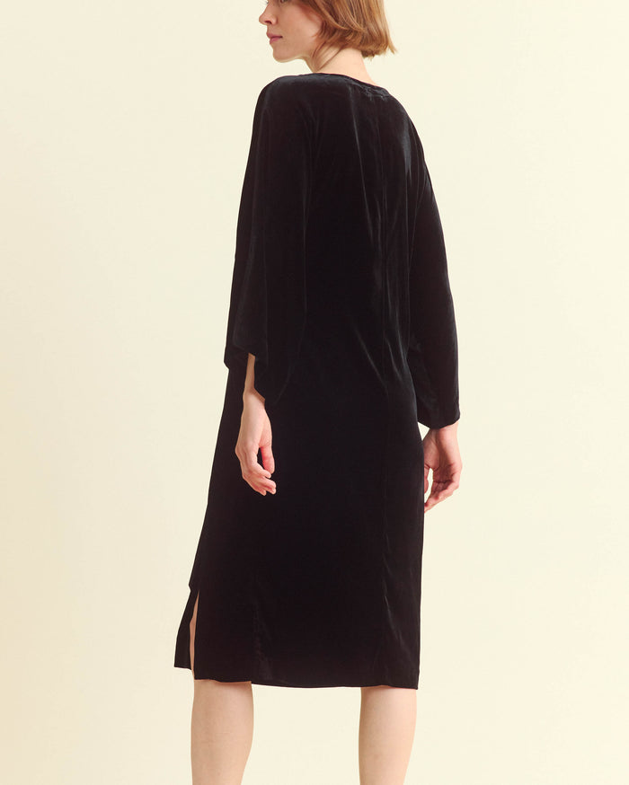 A person with short light brown hair is wearing a loose-fitting, knee-length black velvet dress with three-quarter sleeves and a small slit on the side, shown from the back against a plain background.