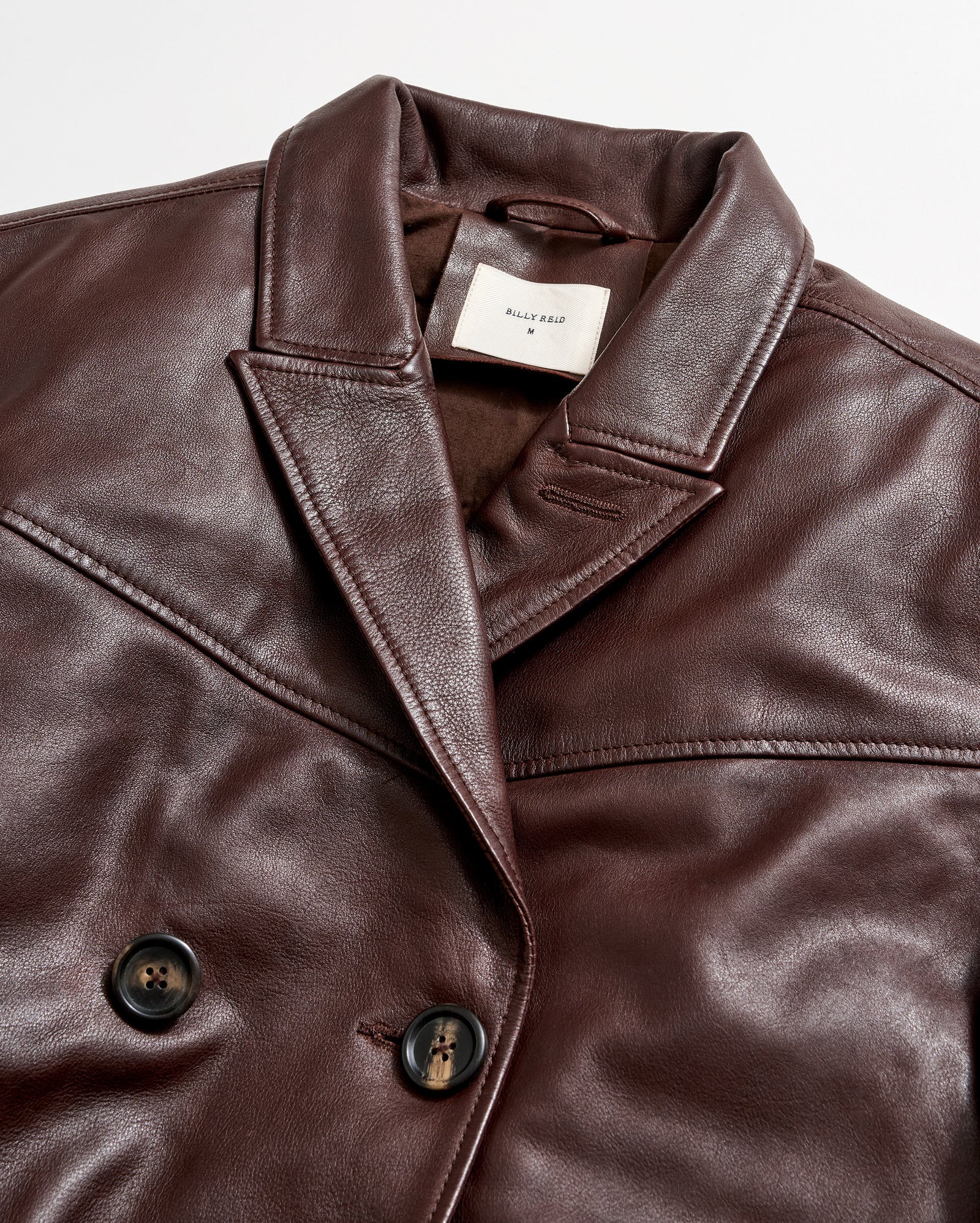 Close-up of a brown leather double-breasted jacket with black buttons and a notched collar. A white label with text is sewn inside the collar. The jacket is laid flat on a white background.