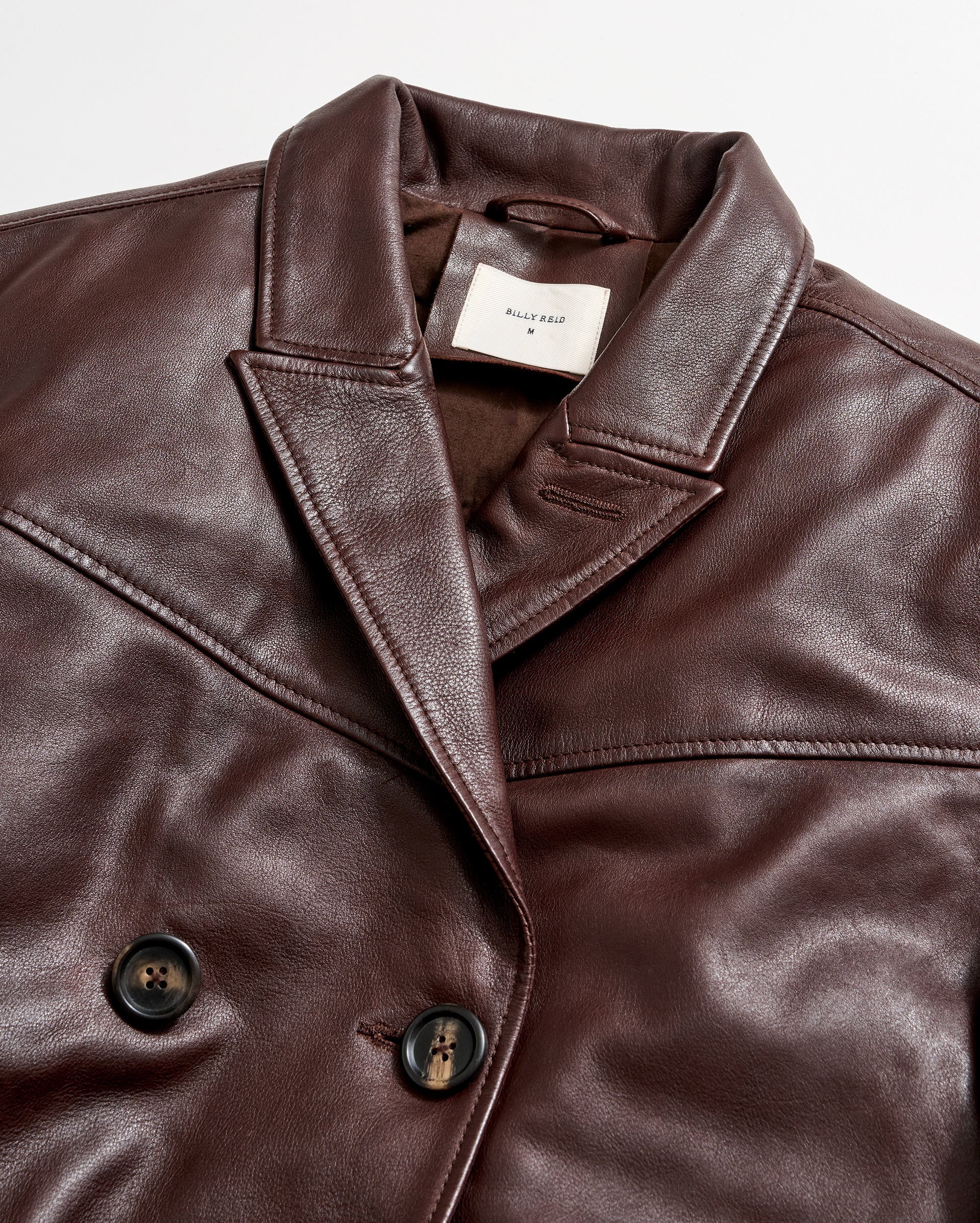 Close-up of a brown leather double-breasted jacket with black buttons and a notched collar. A white label with text is sewn inside the collar. The jacket is laid flat on a white background.