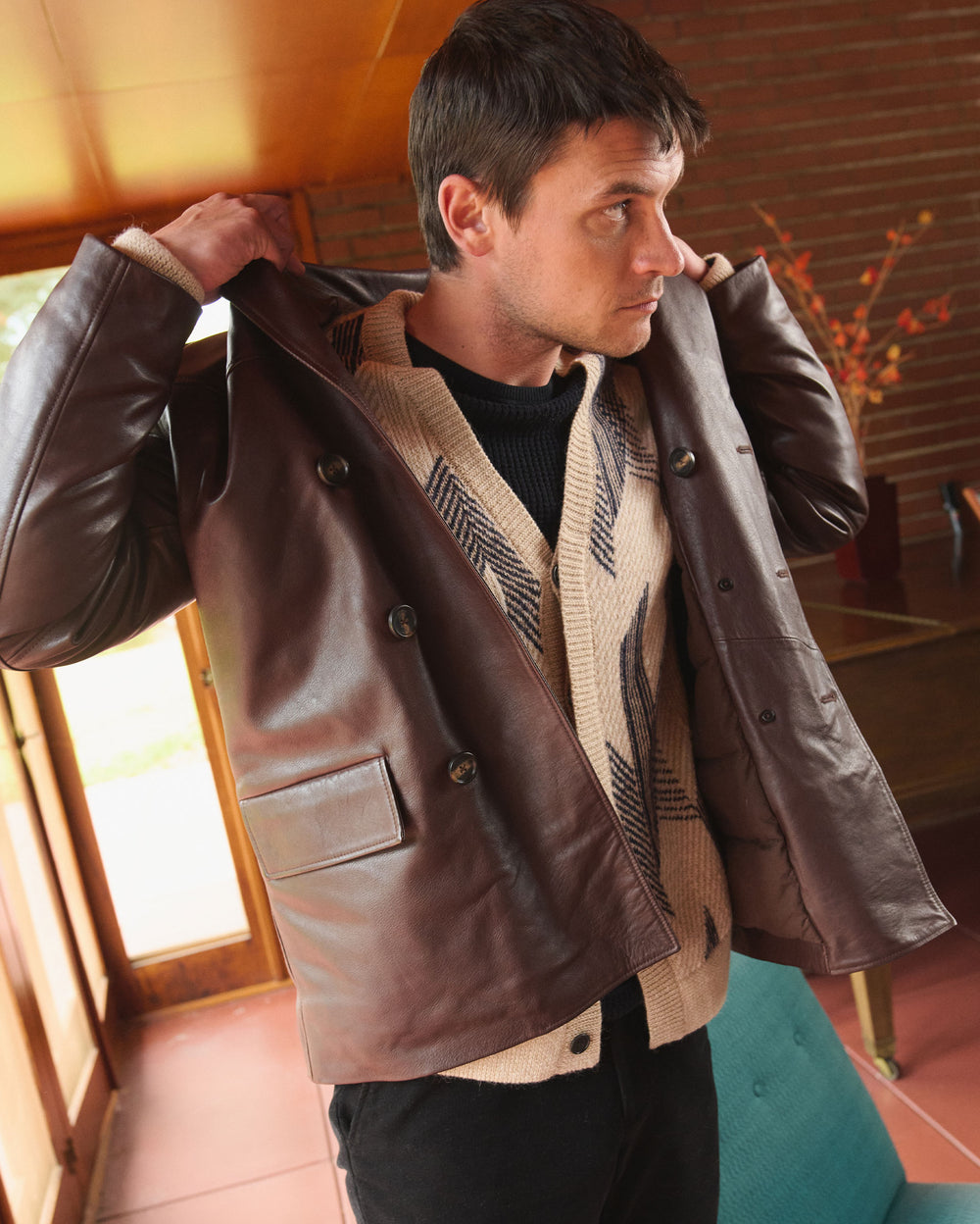 A man standing indoors, lifting a brown leather jacket over his shoulders. He is wearing a beige patterned sweater and black pants, and looking to the side. The room has brick walls and a wooden door.