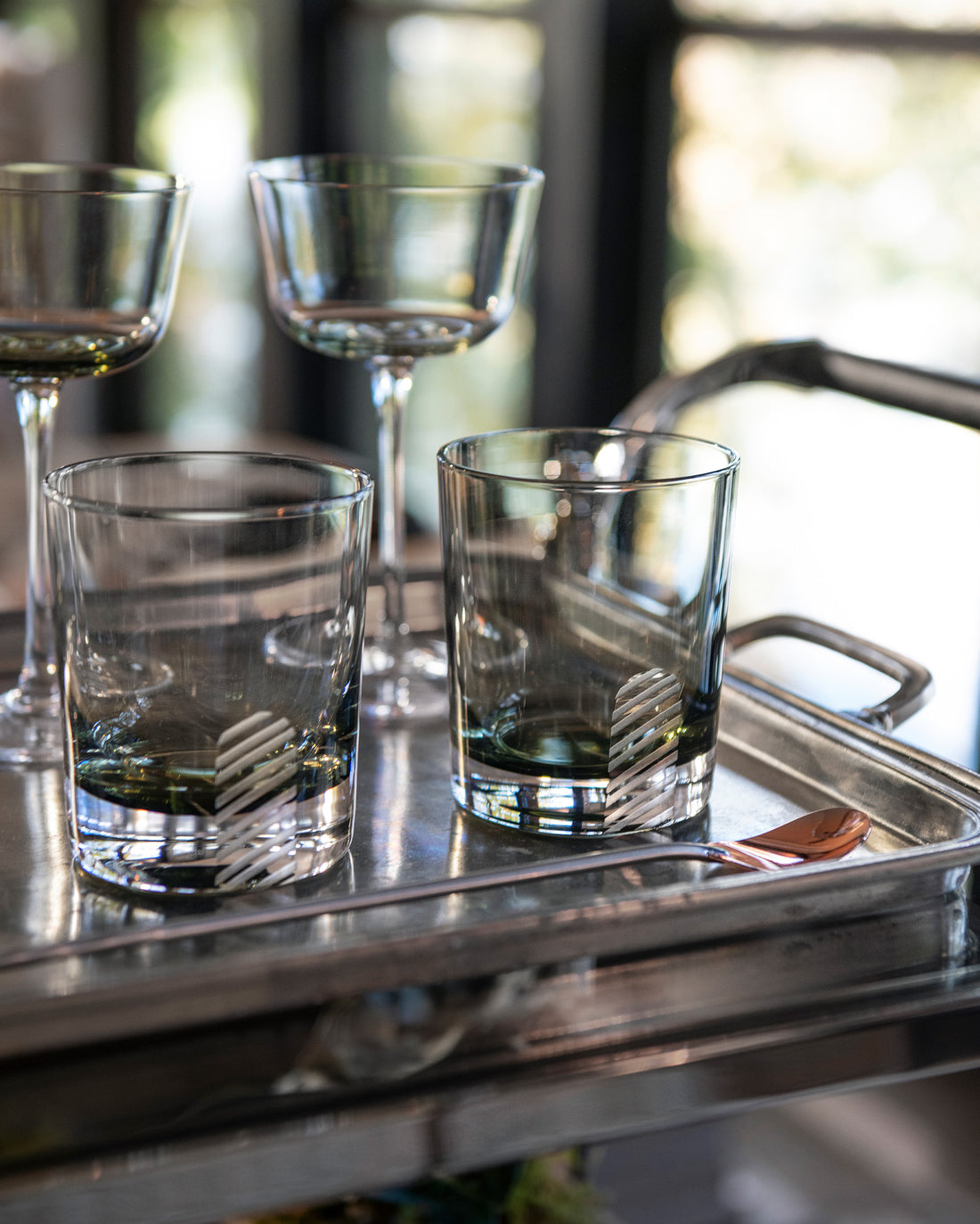 Four empty glasses—two short, two tall—sit on a metal tray with a small copper spoon. The background shows blurred windows and greenery, creating a bright and inviting atmosphere.