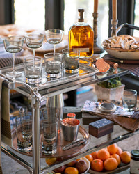 A stylish bar cart with assorted glasses, a bottle of liquor, a bowl of sliced bread, cocktail tools, a potted plant, candles, and a tray of fresh citrus fruits. The background shows a cozy, sunlit room.