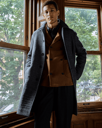 A man stands indoors in front of large wooden-framed windows, wearing a grey overcoat over a brown blazer and black pants. Green trees and buildings are visible outside.
