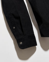 Close-up of a black long-sleeve shirt or overshirt laid flat on a light gray background, showing the sleeve cuff with two buttons and part of the torso area.