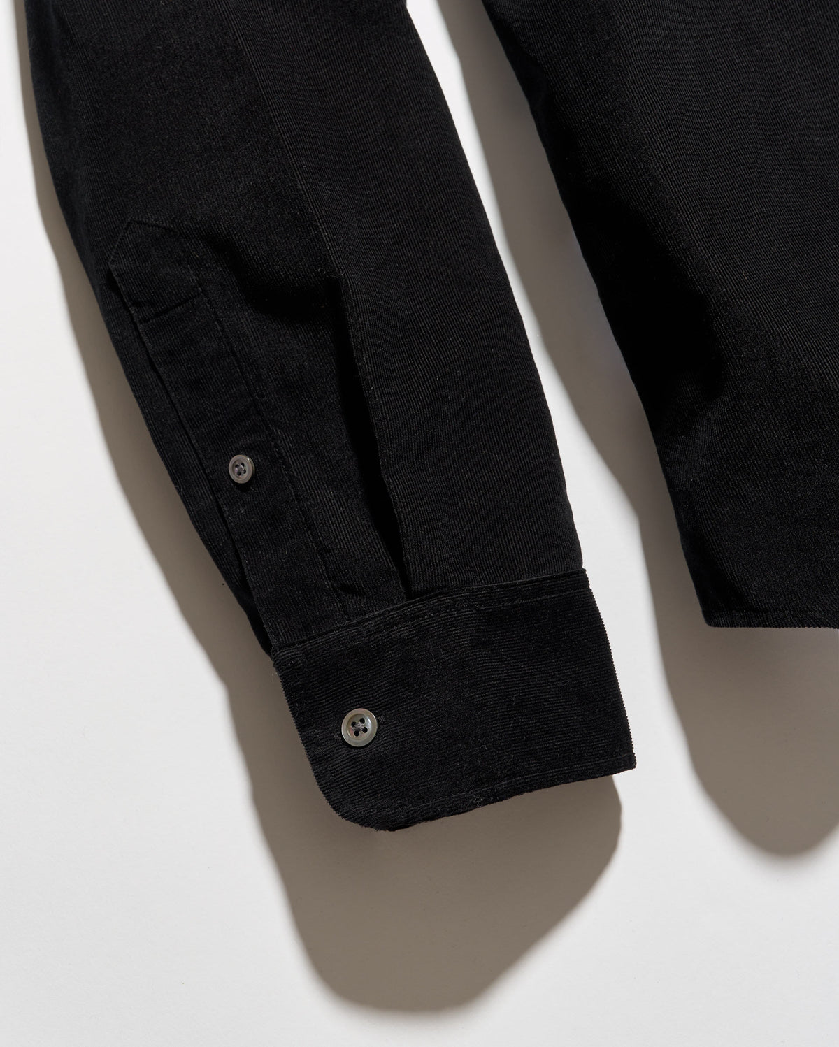 Close-up of a black long-sleeve shirt or overshirt laid flat on a light gray background, showing the sleeve cuff with two buttons and part of the torso area.