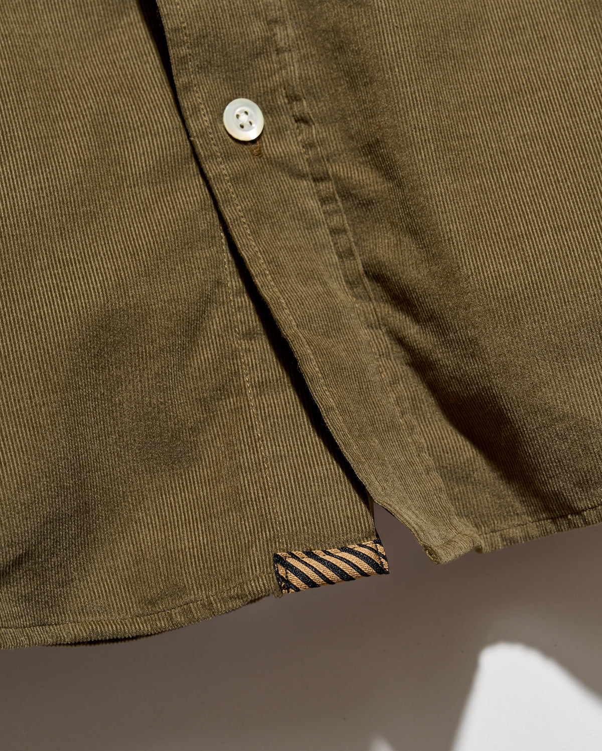 Close-up of an olive green corduroy button-up shirt showing the button, fabric texture, and a striped inner brand tag.