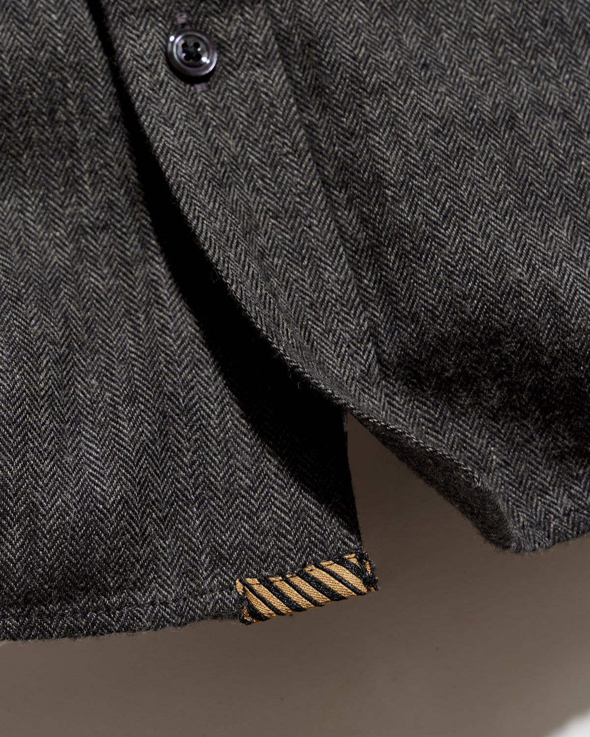 Close-up of a dark gray herringbone tweed jacket with a black button and a gold striped fabric tab near the hem.