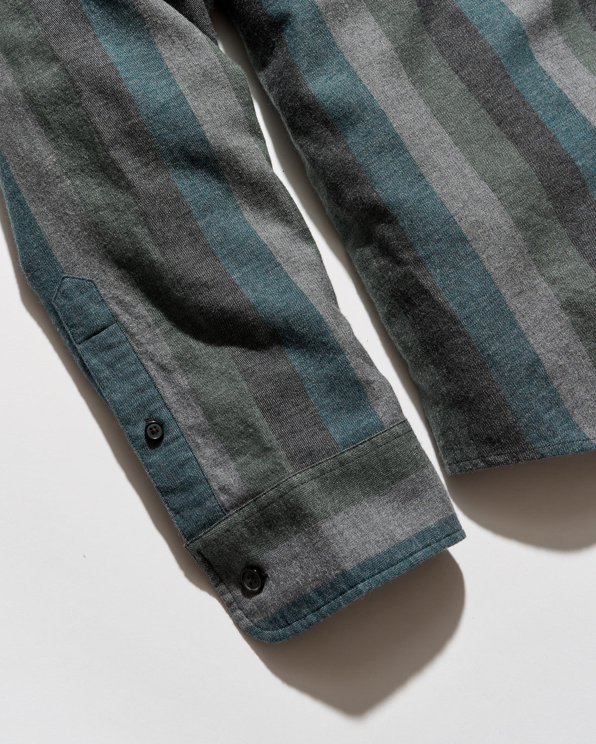 Close-up of a striped shirt sleeve with a teal, gray, and charcoal pattern, featuring a buttoned cuff on a white background.