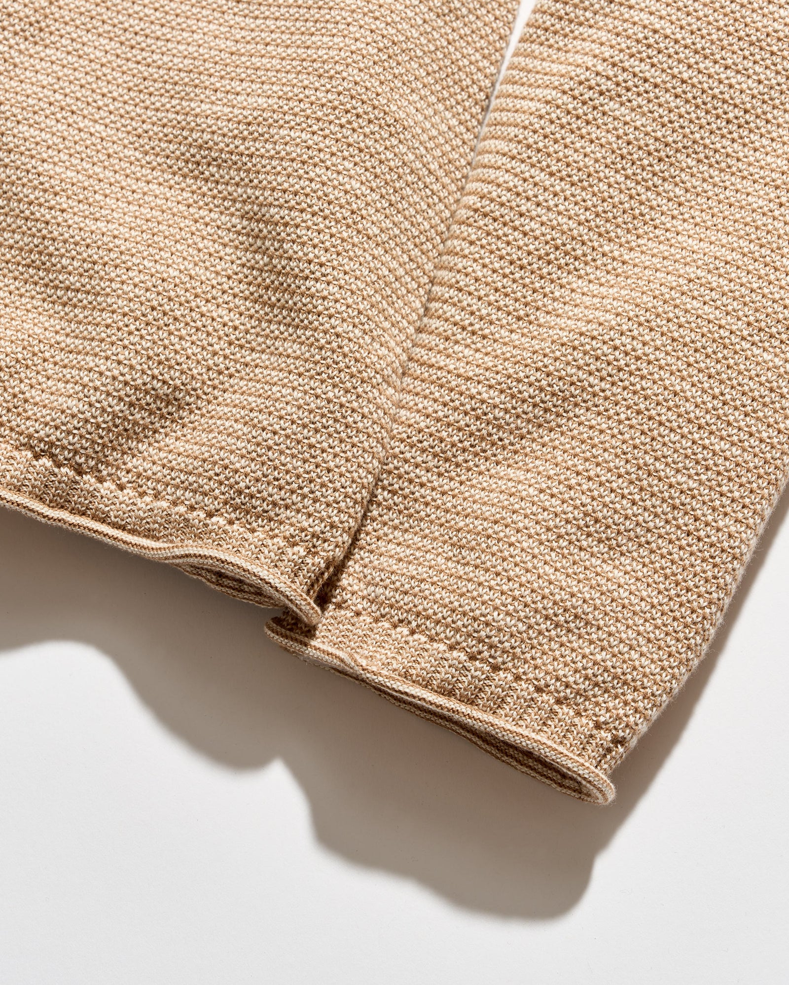 Close-up of a beige knit fabric sleeve or garment detail with visible stitching and a ribbed cuff, photographed on a light neutral background.