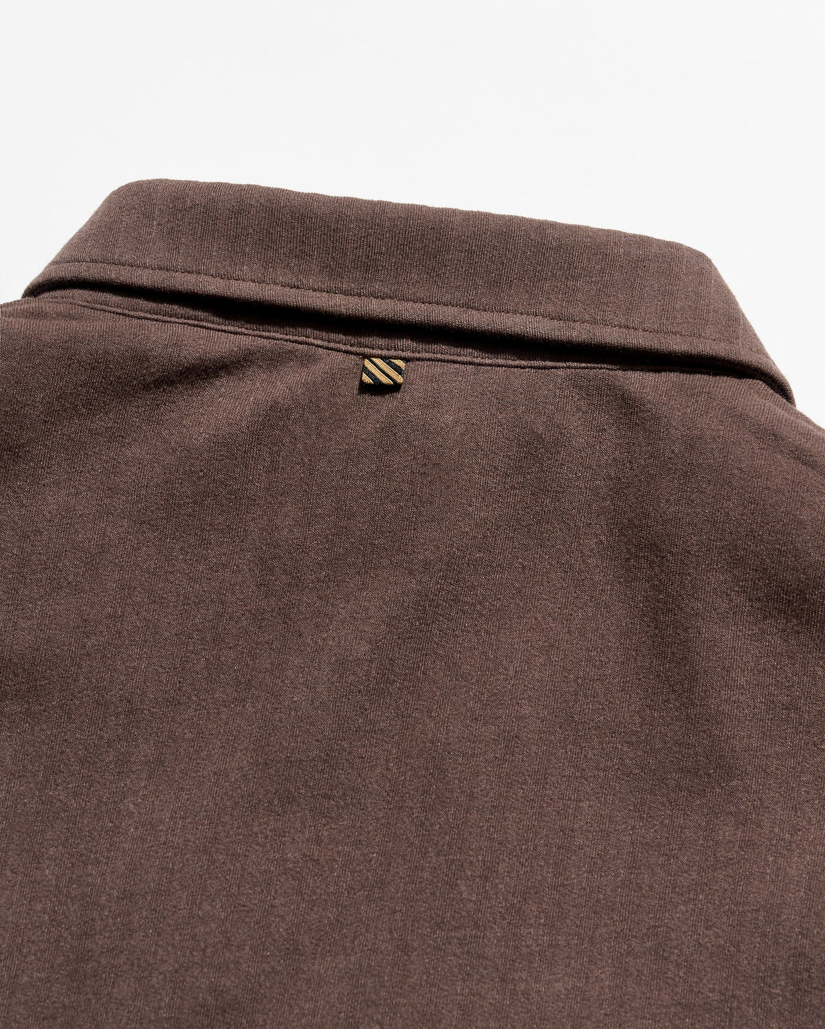Close-up of a dark brown textured fabric garment, showing the back of a collared shirt or sweater with a small striped triangular brand tab near the collar.