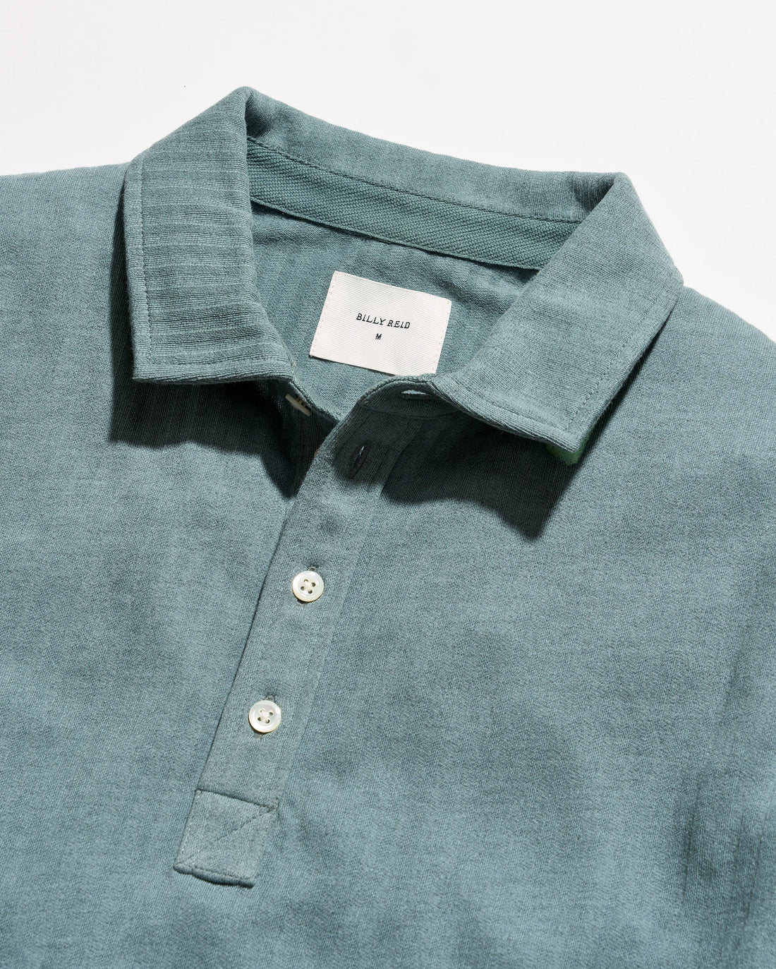 Close-up of a slate blue-green polo shirt by Billy Reid, showing the textured fabric, collar, and white buttons.