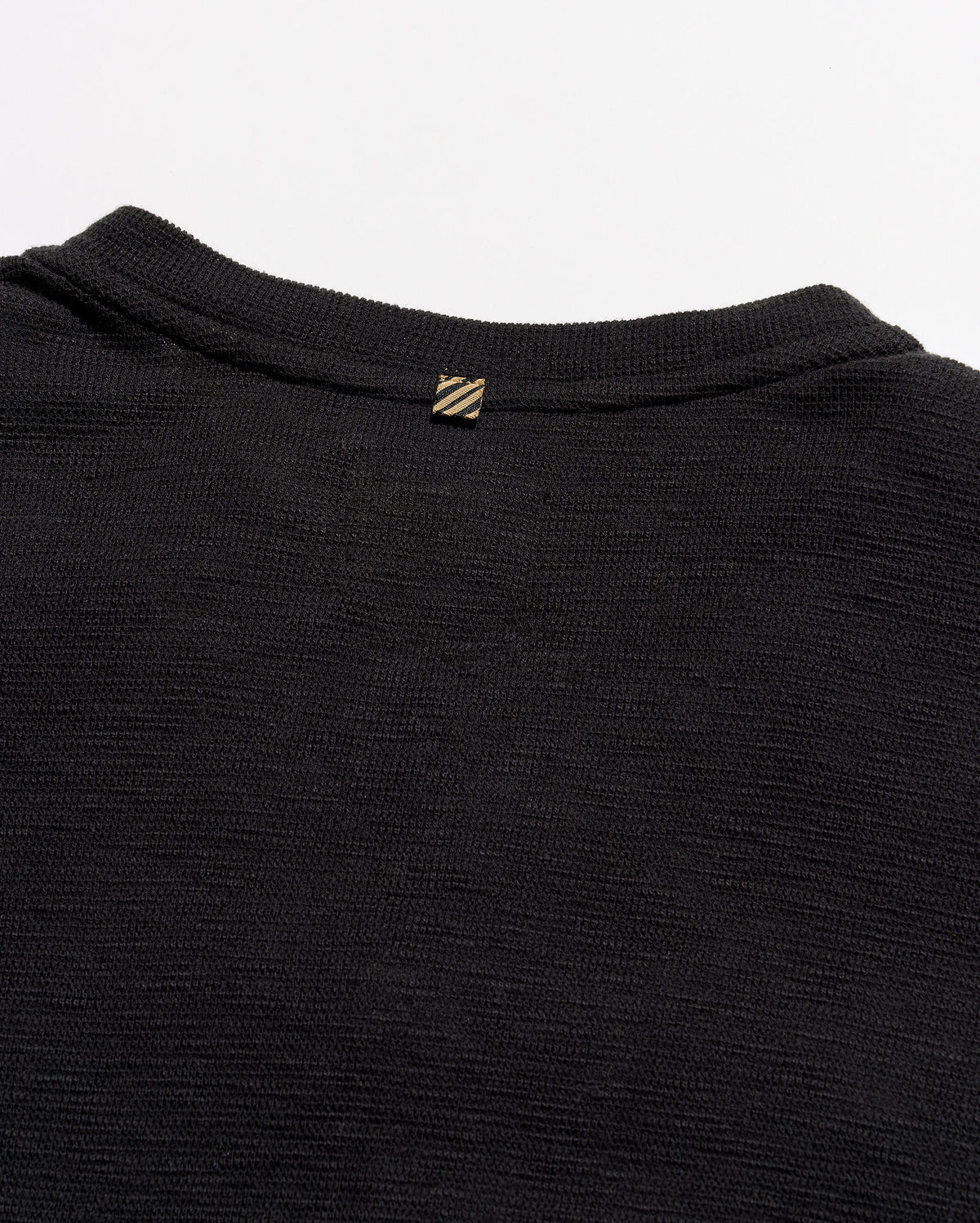 Close-up of the back collar of a black ribbed knit sweater, featuring a small gold-tone embroidered logo with diagonal stripes.
