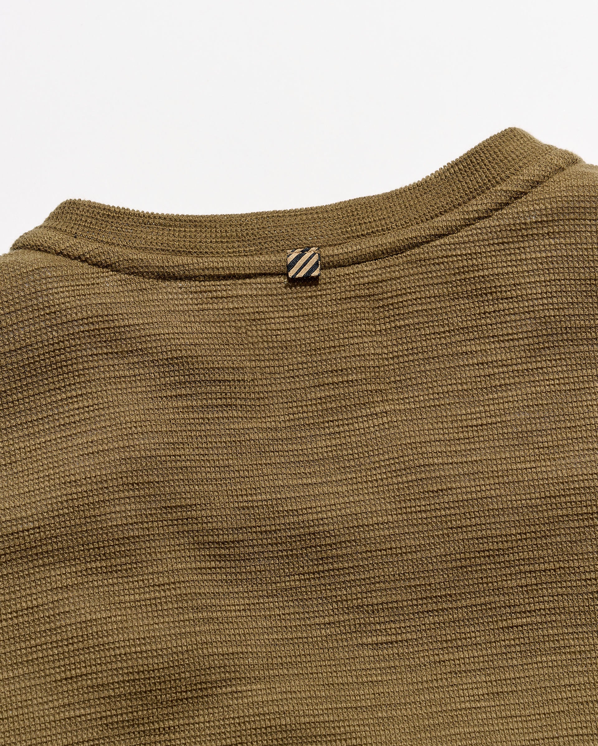 Close-up of a ribbed knit crewneck sweater in olive brown, showing the collar and a small striped button detail against a light gray background.