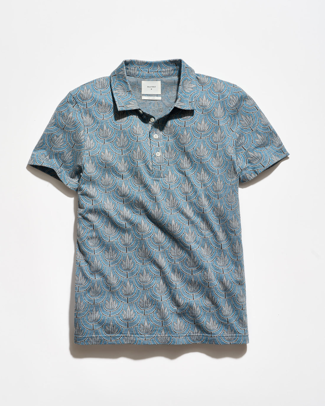 A short-sleeve polo shirt in a dusty blue-gray color, featuring an all-over repeating beige palm leaf pattern. It has a dark collar and a three-button placket, laid flat against a light off-white background.